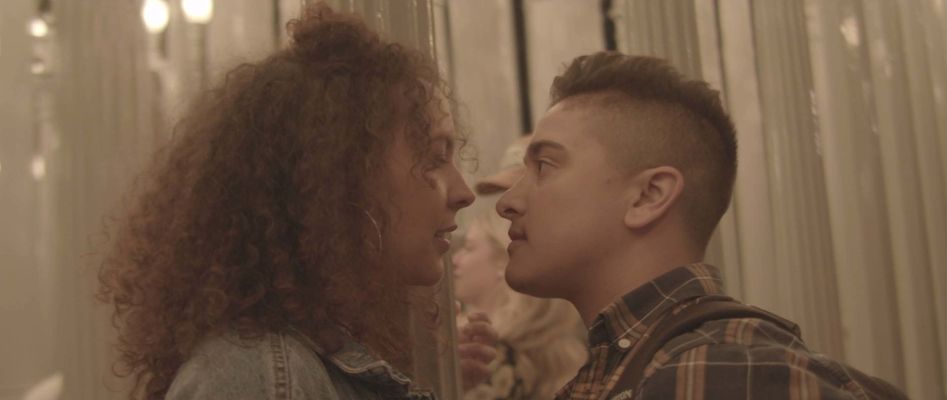 Two people stand really close together facing each other and the background is blurred out white street lamps. The person on the left has tan skin, shoulder length curly hair, wearing hoop earrings and a denim jacket. The person on the right has tan skin, short brown hair with the sides faded, wearing a flannel t shirt.