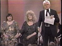 Still of three people in a talk show environment; two seated women and a standing male with white hair holding a microphone and paper. The seated women are holding hands. The one on the left has hair that is black and extremely straight, while the one on the right has blonde hair that is extremely curly. The heads of an audience are visible on the bottom of the frame.