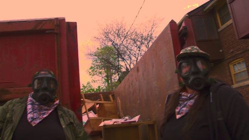 Two people standing wearing gas masks in front of a house and a dumpster looking at the camera. The one on the left is wearing a camouflage flannel and a black t-shirt and a pink and blue patterned bandana around their neck. The person on the right has the same bandana and they are wearing a black zip up with a black t-shirt underneath.