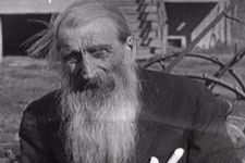 An archival black and white photograph of an elderly man in the the Canadian prairies, circa 1920-1940. The man has long light hair and a long light beard, and he wears a black coat. The stairs of a homestead are visible in the background.