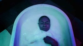 A person lays in a white tub with only their face and right hand sticking out of the white goo they are lying in. They have dark brown skin with their eyes closed and they have a buzz cut.
