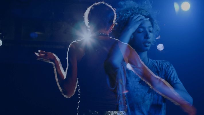 A person is standing with their back to their camera and a person is facing the camera but they are more transparent with an image overlaid on top of the original image. The overlaid image shows a person with light brown skin and long curly hair pulled into an updo. They are resting their left hand on their forehead and wearing a gray short sleeve shirt. The person with their back to camera is wearing a dark-colored tank top, a bunch of silver necklaces stacked, and a silver glitter belt. They have light skin and short brown hair. The background is black with blue lights shining on the two people.