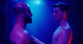 Two shirtless people stand in front of each other and stare intently at one another. The person on the right has light skin and short brown hair with the sides faded. They are touching the chest of the person on the left with their left hand. The person on the left has light skin with a beard and a hairy chest wearing a thin gold necklace.