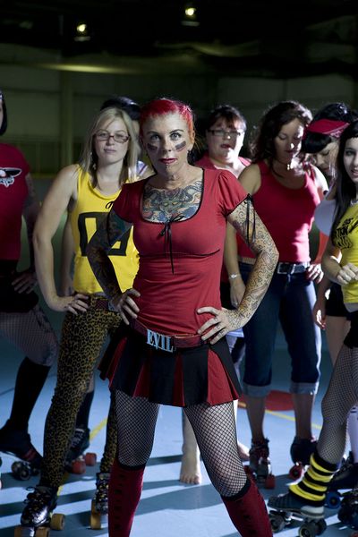Eight people pose with one person in the forefront. The person in the front has light skin, tattoos covering their chest and arms. They have a red short sleeve t-shirt, a red and black pleated skirt, a red belt with the belt buckle that says "evil", and knee high socks. They are posed with their hands on their hips, they have red hair pulled back, a piercing under their lip, and red and black striped face paint on either cheek. The person behind them to the left is posed in the same way with light skin, blonde hair and glasses. They are wearing a yellow tank top and yellow cheetah print pants with roller skates on. There are varying people behind them posing, some with red shirts and some in yellow, most in roller skates. The background is dimly lit with the focal point of the light on the group. The floor is blue with yellow lines on the floor and some red circles.