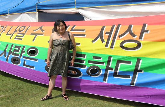 A person stands on grass in front of the pride flag with Korean writing on it. They are wearing a strapless dark green dress. Their arms are relaxed by their sides with a black purse on their right arm. They are wearing a silver pendant necklace with a black string and black sandals. They have a bob cut of black hair and parted bangs and they are smiling at the camera.