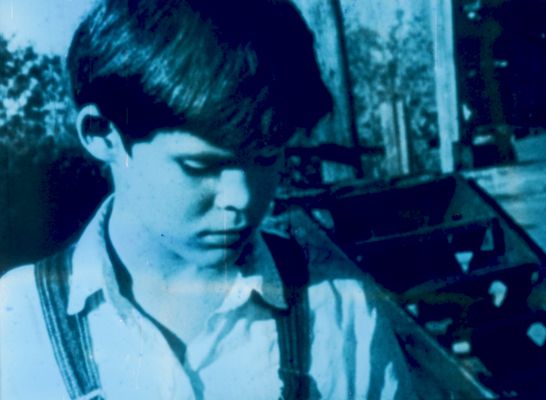 A close up of a light-skinned child gazing toward the ground. They appear from the shoulders up, have short dark hair, and are wearing a collared shirt with overalls. They are outside in front of bushes and a staircase.