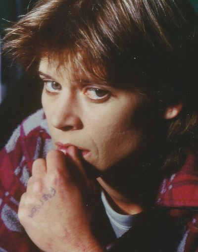 Brooke Johnson as Marlene Moore in Dangerous Offender. They look into the camera with green eyes, light skin, short parted down the middle brown hair, and wearing a red and white patterned sweater with a white t-shirt underneath. They are resting their right hand on their lip and a few letters written on the back of their hand that are not legible.
