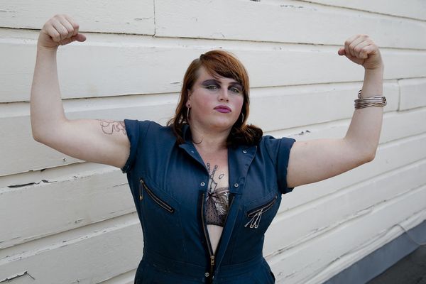 A person is posing with both of their arms flexing in an L position towards the camera. They wear a denim short-sleeve jumpsuit with the front unzipped, displaying their black-lace bra. They have straight light brown shoulder-length hair. They are wearing purple eyeshadow and pink lipstick, and silver bracelets on their right wrist. A tattoo is partially visible on their left upper arm, and another one is partially visible on their chest.