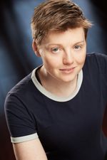 Studio headshot of Amy Fox, a white transgender woman with tousled short brown hair. Amy wears a navy t-shirt with white lining, and stares up at the camera with a defiant expression.