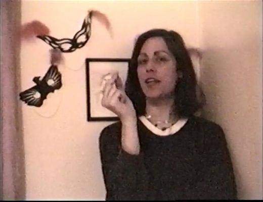 Xanthra Phillipa Mackay, a white trans woman with shoulder-length dark hair, sits in a home setting, speaking to the camera. Behind her are two decorated, feathered masks, likely for theatrical or party use.