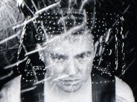 A creative self-portrait taken by Malic Amalya. Malic Amalya has fair skin, short dark hair, and is wearing a black tank top. They look towards the camera pointed above with their eyes only, posing with a serious and focused expression. The image is a black-and-white photograph with a surreal and intense atmosphere created by eerie lighting, the dark background, superimposed metallic chain elements and abstract fragmented patterns overtop the portrait.