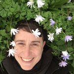 Coral Short, a nonbinary person with light skin and short dark hair, is lying on a bed of green leaves and flowers. They are smiling brightly at the camera, surrounded by small white and light purple flowers.