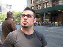 Joe Ippolito is standing on a bustling city sidewalk, wearing a black t-shirt and black-framed glasses. Joe has short dark hair, a goatee, and is photographed in action while focusing on something in the environment behind and above the camera.