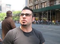 Joe Ippolito is standing on a bustling city sidewalk, wearing a black t-shirt and black-framed glasses. Joe has short dark hair, a goatee, and is photographed in action while focusing on something in the environment behind and above the camera.