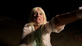 Still of Dominique Dibbell in a moment of dramatic expression. She is a white person with blonde hair and pistachio-green eyeshadow. They are wearing a white jacket over a lacy seafoam-green shirt, with a long pearl necklace draped about their neck. Her arm is outstretched in an emphatic gesture, with eyes wide and teeth bared in apparent terror.
