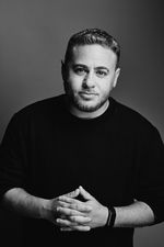 Black-and-white professional studio portrait of Alex Schmider. Alex has light skin, a short tapered hairstyle, and facial hair. He is wearing a dark long sleeve shirt and a wrist watch.