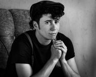 Vintage style black-and-white photograph of Kai Tillman posing with bent arms on an armchair. Kai has fair skin and dark, curly ear-length hair with sideburns. He is wearing a dark flat cap, a dark t shirt and vintage patterned suspenders.
