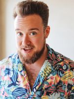 Bright and summery portrait of Zeke Smith with slightly widened, animated eyes and open smile. Zeke has light, tanned skin, medium short light brown hair that is styled upward, and light brown facial hair. He is wearing a colourful hawaiian shirt with chest hair exposed.