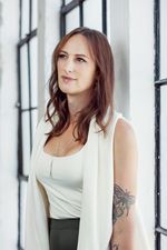 Bright and airy professional portrait of Jen Richards in a white loft studio with black industrial windows. She has fair skin and auburn middle-parted shoulder length hair that enhances her brown eyes. She is wearing a white shirt and sleeveless cardigan with black bottoms. A colourful butterfly tattoo is visible on her left upper arm.