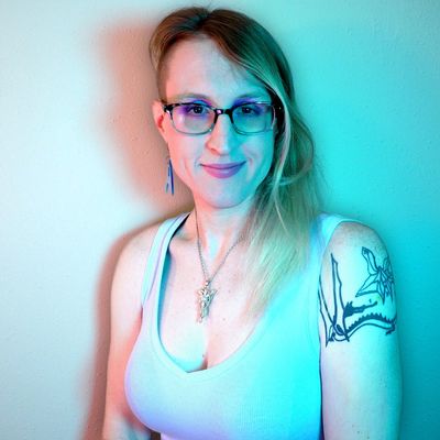A straight-on photograph of Jessie Earl in front of a plain wall with interesting cyan lighting colouring the right side of the potrait. Jessie has fair skin, medium-length blonde hair with one side shaved.
