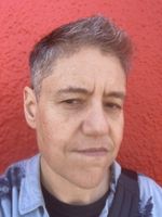 Jed Bell gives the camera a wry sideways glance in front of a red wall in this punky selfie. Jed is a trans man with light skin and spiky gray-black hair. He wears a light denim shirt.