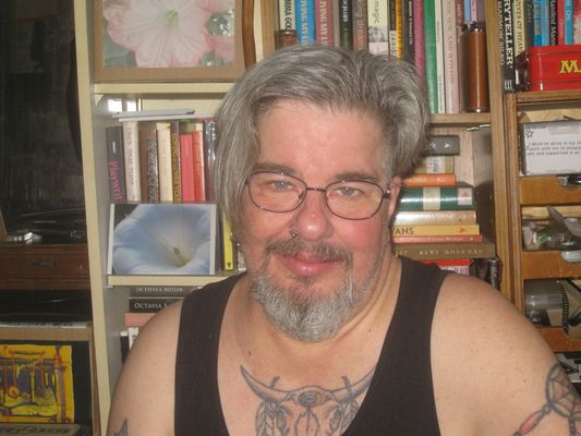AAlec Whitewolf Butler looks at the camera with a slight smile in front of a bookcase. They are a Two-spirit Mi’kmaq person with light skin, grey hair, light eyes, and a beard. They wear metal-rimmed glasses, a tank top and have tattoos.