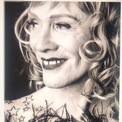 Close cropped black-and-white photo of Karla Rae James, who has light skin and light curly hair to her shoulder. She wears lipstick, has star tatoos on her shoulder and a few feathers likely from an outfit just outside the frame.