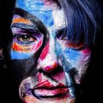 Close-up of Brandubh's face covered in a painterly and expressive face paint. One eye stares at the camera while the other is obscured by blue hair falling in front of their face.