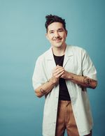 Amos Mac stands in front of a dark eggshell-blue background and stares into the camera, his fist in his hand. He has light skin, a mustache, and a stylish wave of dark hair atop his head. He wears a white labcoat over a plain black shirt and chestnut-brown jeans.