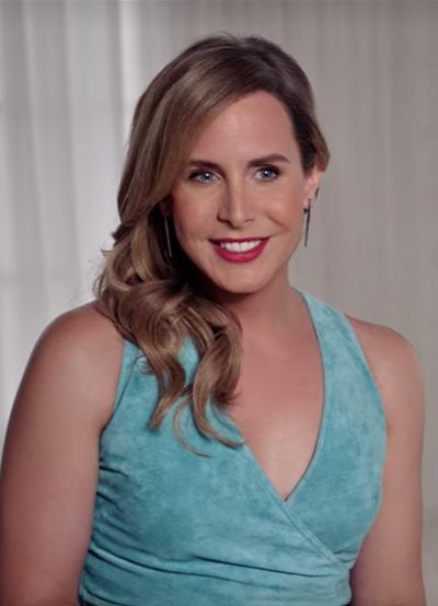 Zackary Drucker smiles past the camera in front of white curtains. Zackary is a trans woman with light skin and ashy blonde hair. She wears red lipstick, dangling earrings, and a light blue dress with a felt-like texture.