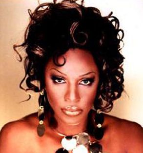 Headshot of Jazzmun, a Black trans woman with curls piled into an updo and glossy lips. She wears statement earings and a necklace of interlocking silver circles.