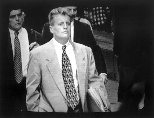 Black and white film still of Leslie Feinberg walking down the street. Ze has light skin and short hair. Ze wears a suit and holds a newspaper under their arm.