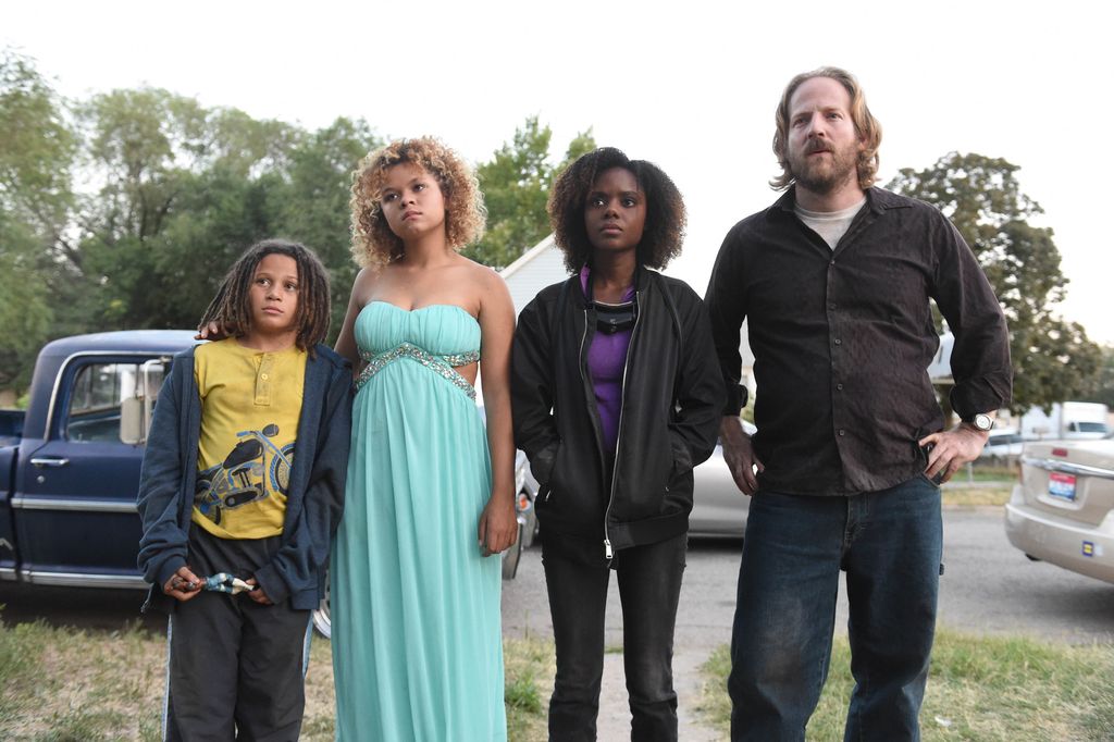 Still from Deidra & Laney Rob a Train (2017). Four characters stand in a line looking incredulously at something behind the camera. The person on the far left is a child with light brown skin and shoulder length dreads. They are wearing a white t-shirt with a black motorcycle on it, a dark blue cardigan, light black pants holding something in their hands. The person to the right of them has light brown skin and blonde curly shoulder length hair with brown roots. They are wearing a strapless light blue dress with silver trimming on the sternum. The person next to them has their hand in their jacket pocket, dark brown skin, with shoulder length dark brown curly hair.They are wearing a black zip up jacket, a purple t-shirt underneath, and black jean pants. The last person to their right is standing with their hands on their hips, light skin, and short straight blonde hair and a small beard. They are wearing a black button up t-shirt with a white undershirt, blue denim jeans and a watch on their right wrist. 