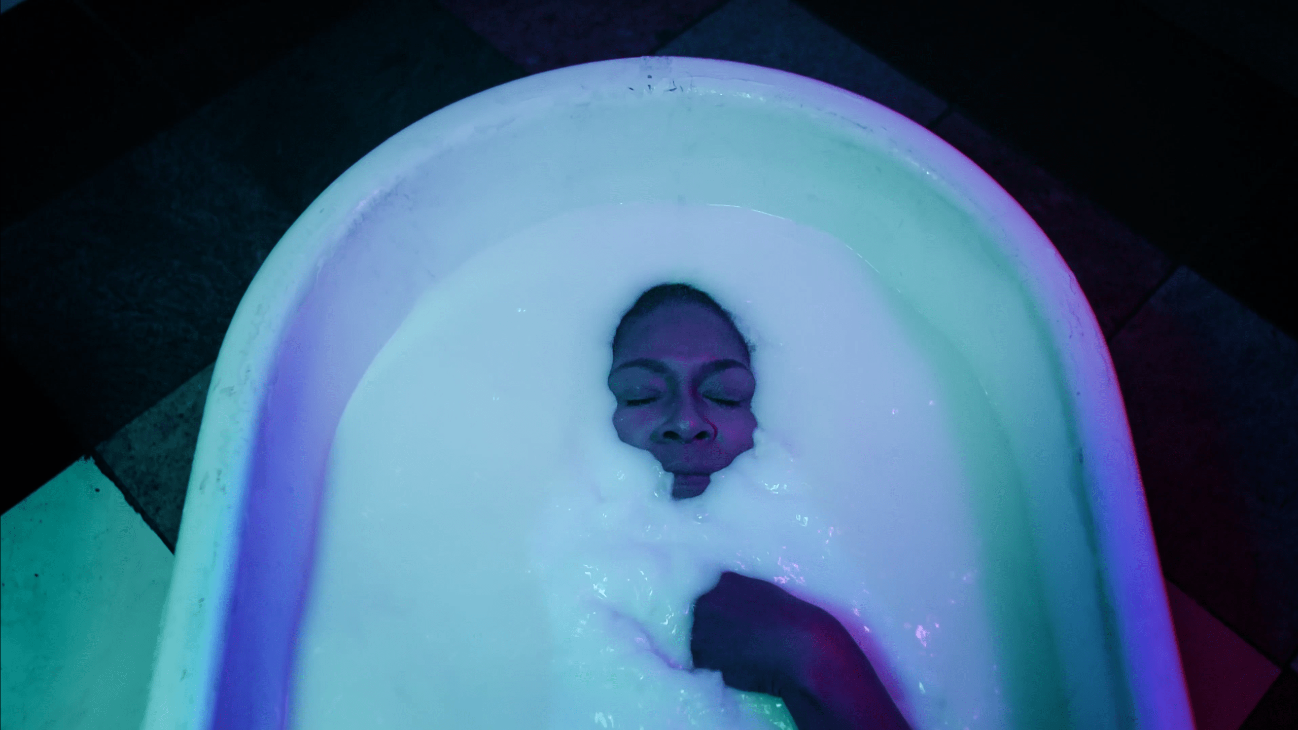 A person lays in a white tub with only their face and right hand sticking out of the white goo they are lying in. They have dark brown skin with their eyes closed and they have a buzz cut.