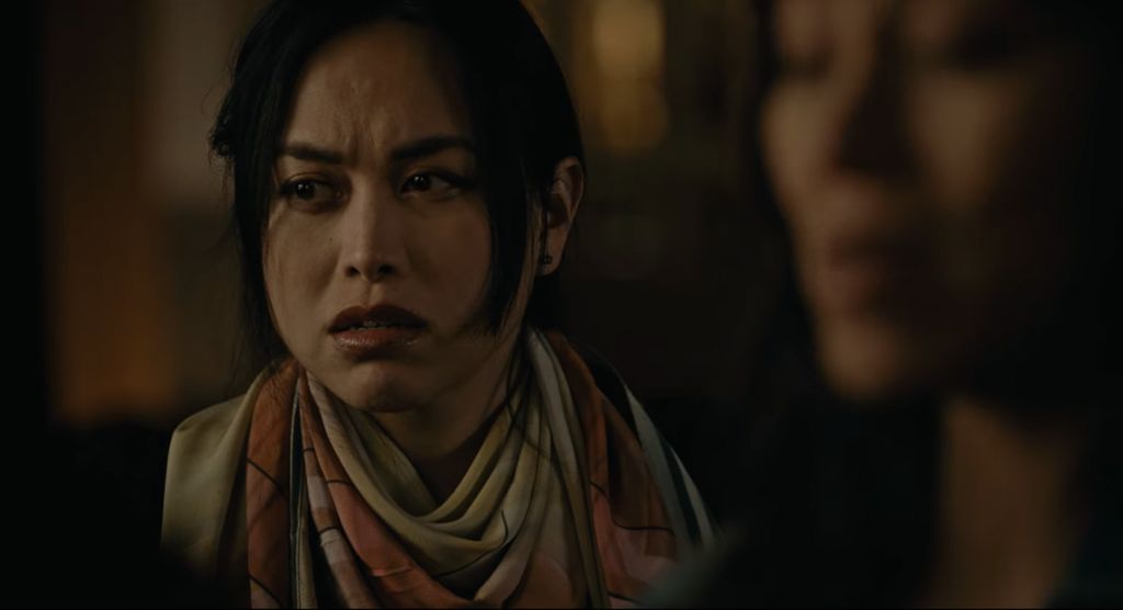 Still from Lingua Franca (2019). A portrait shot of Isabel Sandoval, who has long dark hair pulled back with two strands on either side framing her face. She has light skin and is wearing a tan and burnt orange scarf wrapped a few times around her neck. She is staring at something off camera with her brows slightly furrowed. The shot is  dimly lit with the background blurred out, the focal point being Isabel.